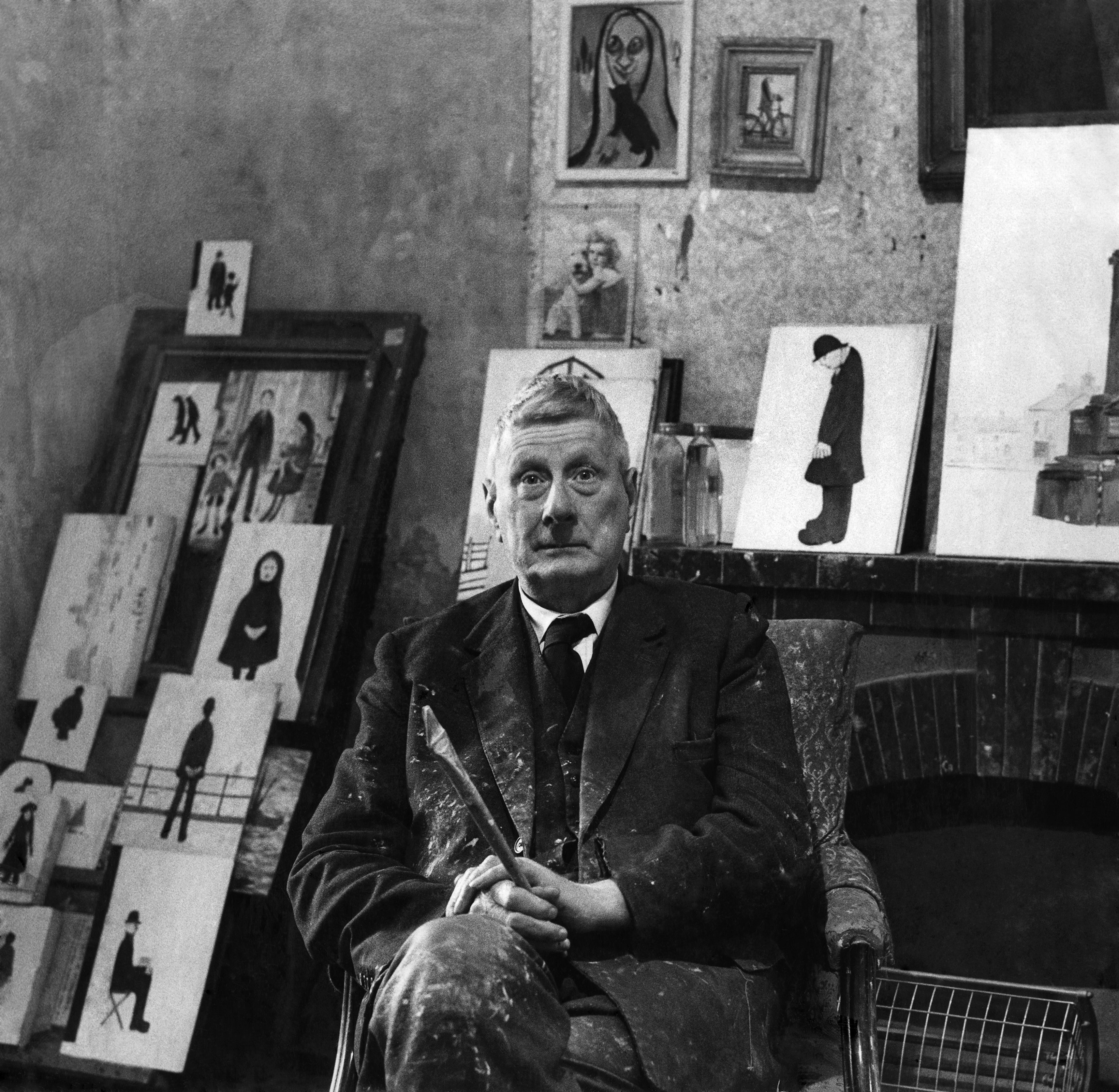 L.S. Lowry sitting in a chair, surrounded by his paintings.