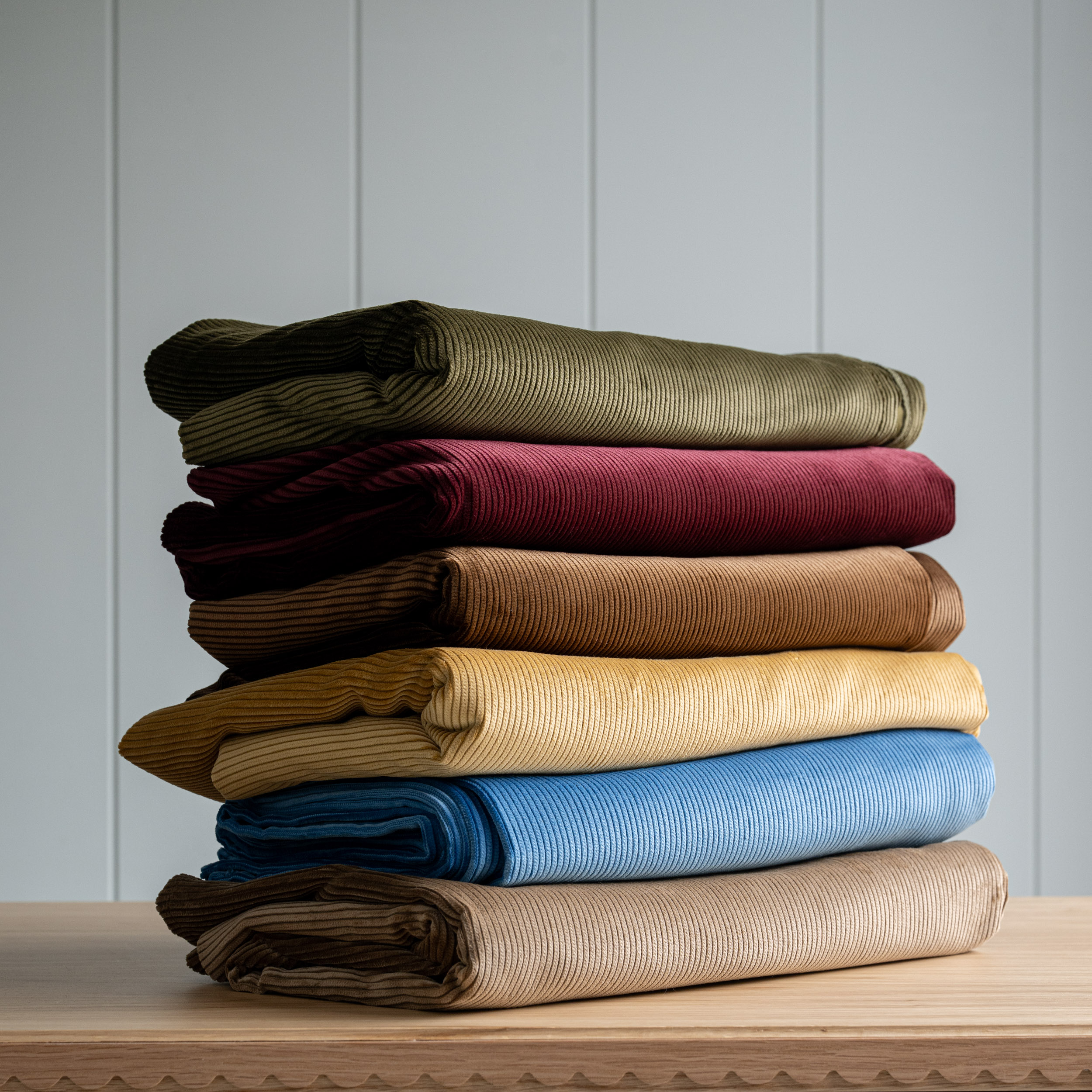 A stack of six pieces of folded corduroy fabric in olive green, dark red, brown, yellow, light blue, and beige.
