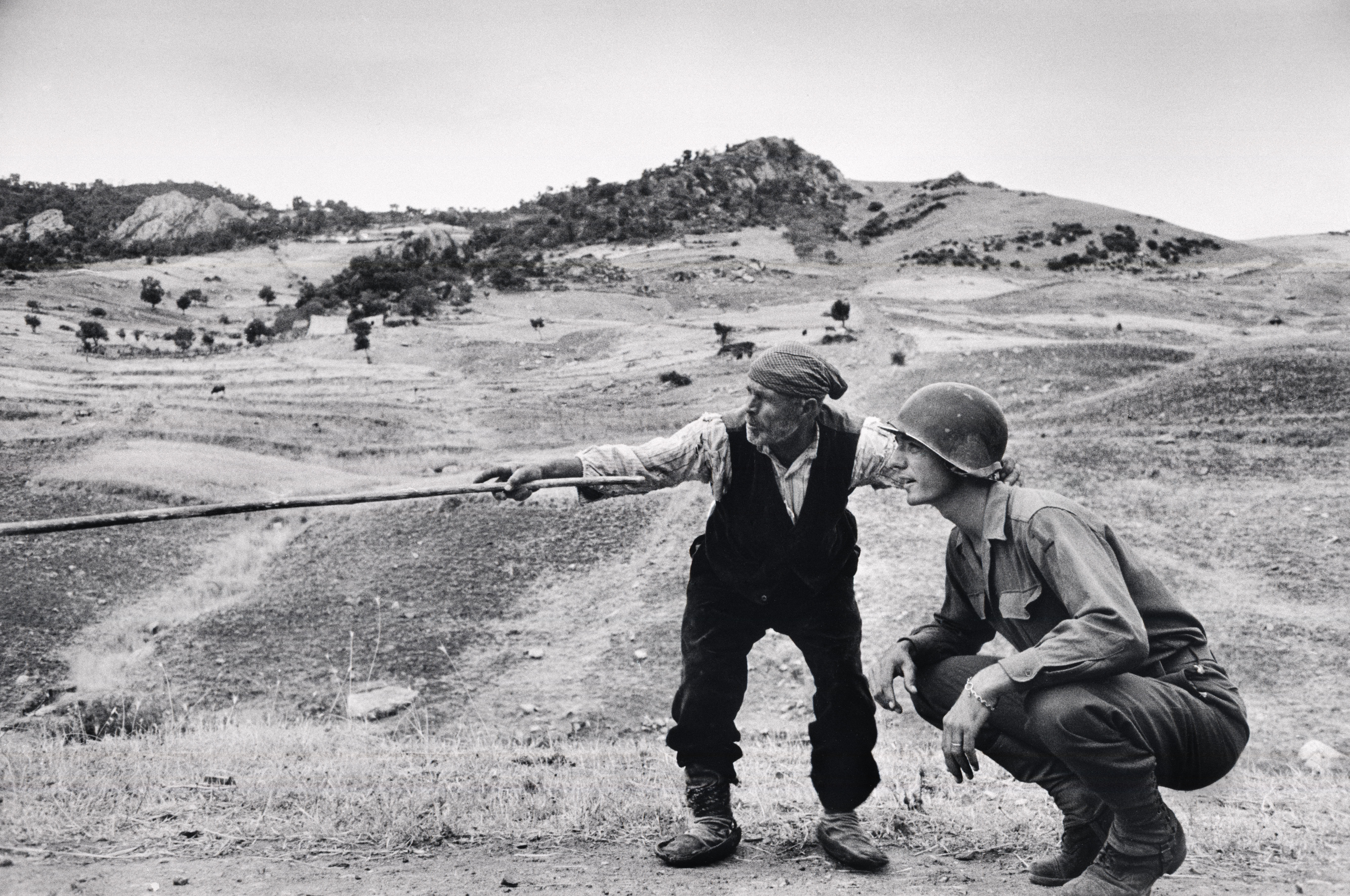 A Sicilian peasant points with a stick to an American officer, who is squatting and listening, in a hilly landscape.