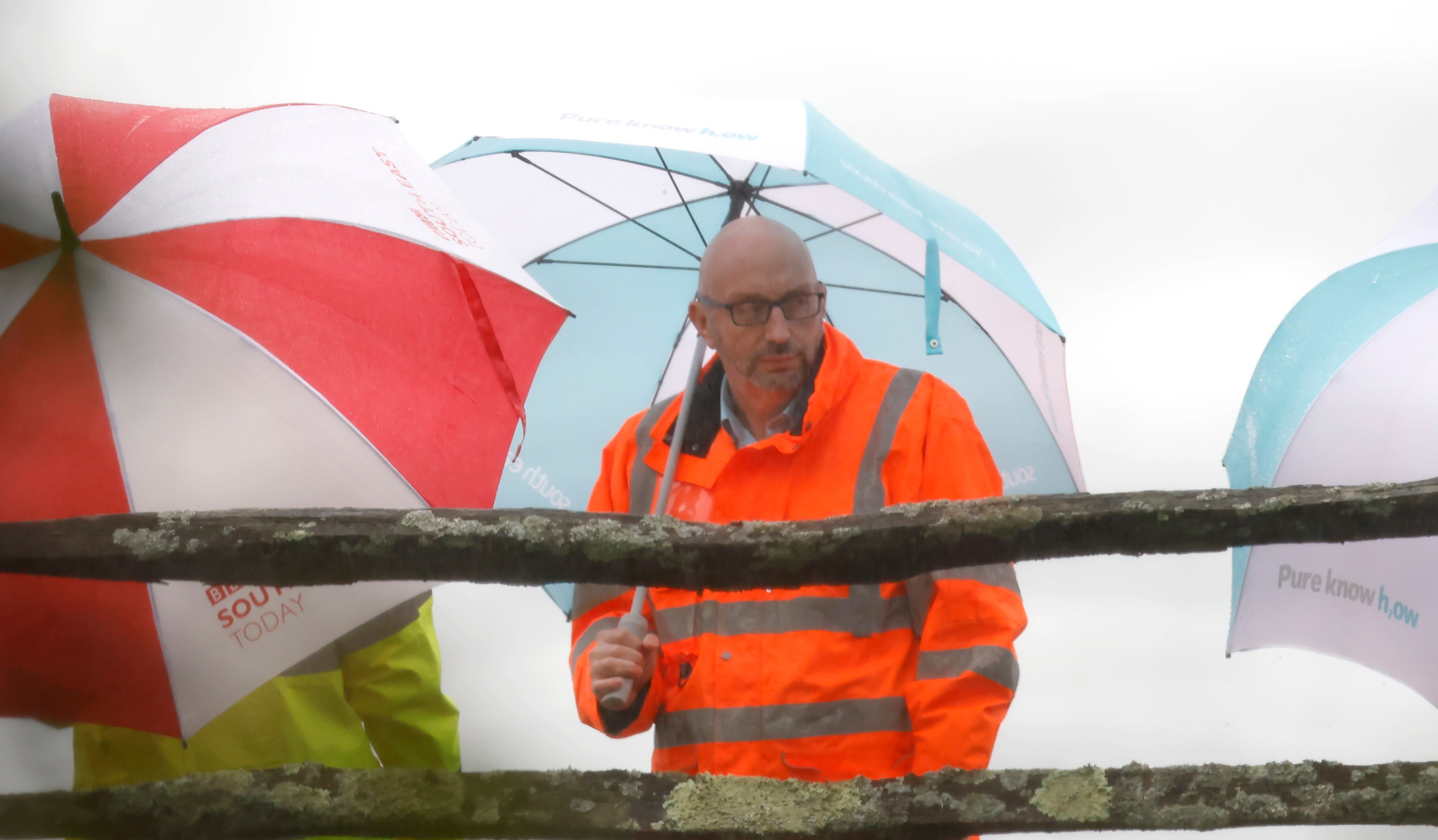 Dave Hinton, CEO of South East Water, seen through a fence, holding a blue and white umbrella while wearing an orange reflective jacket.