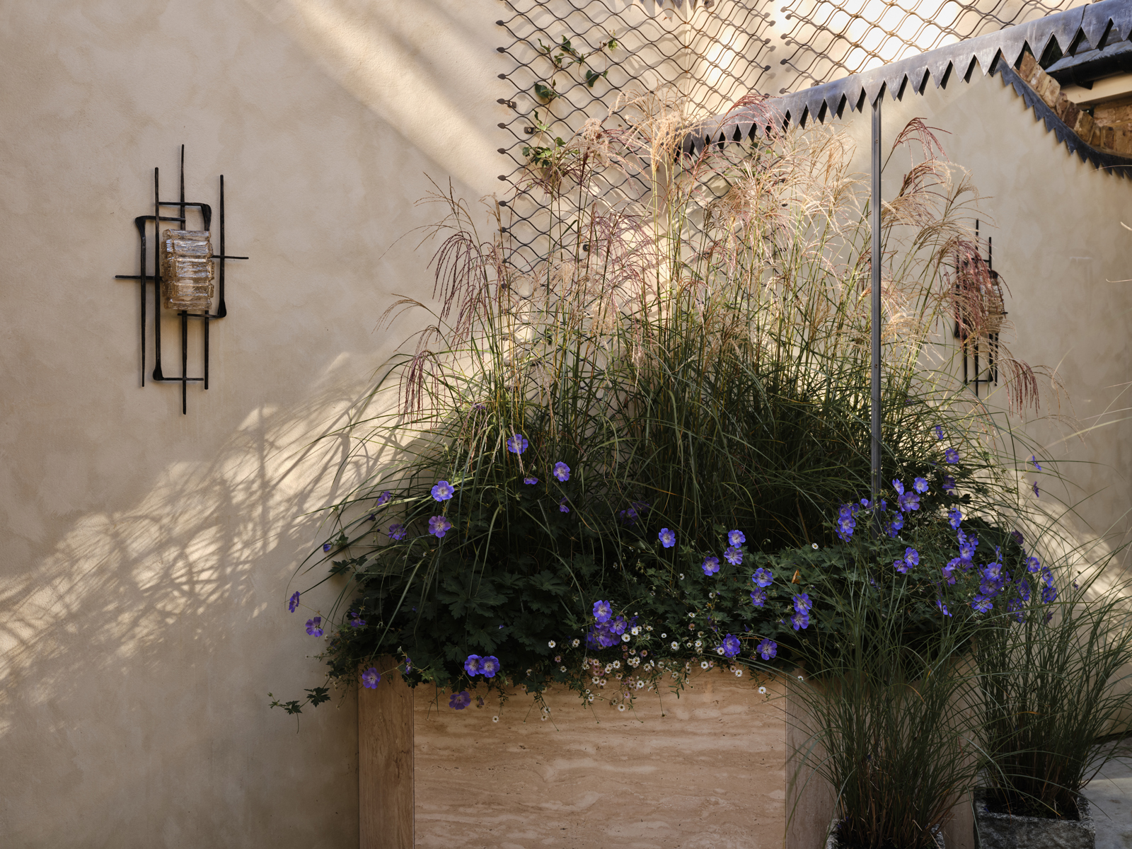 A planter box with tall grasses and purple flowers sits against a cream wall, with a sconce on the left and a decorative metal grid on the right.