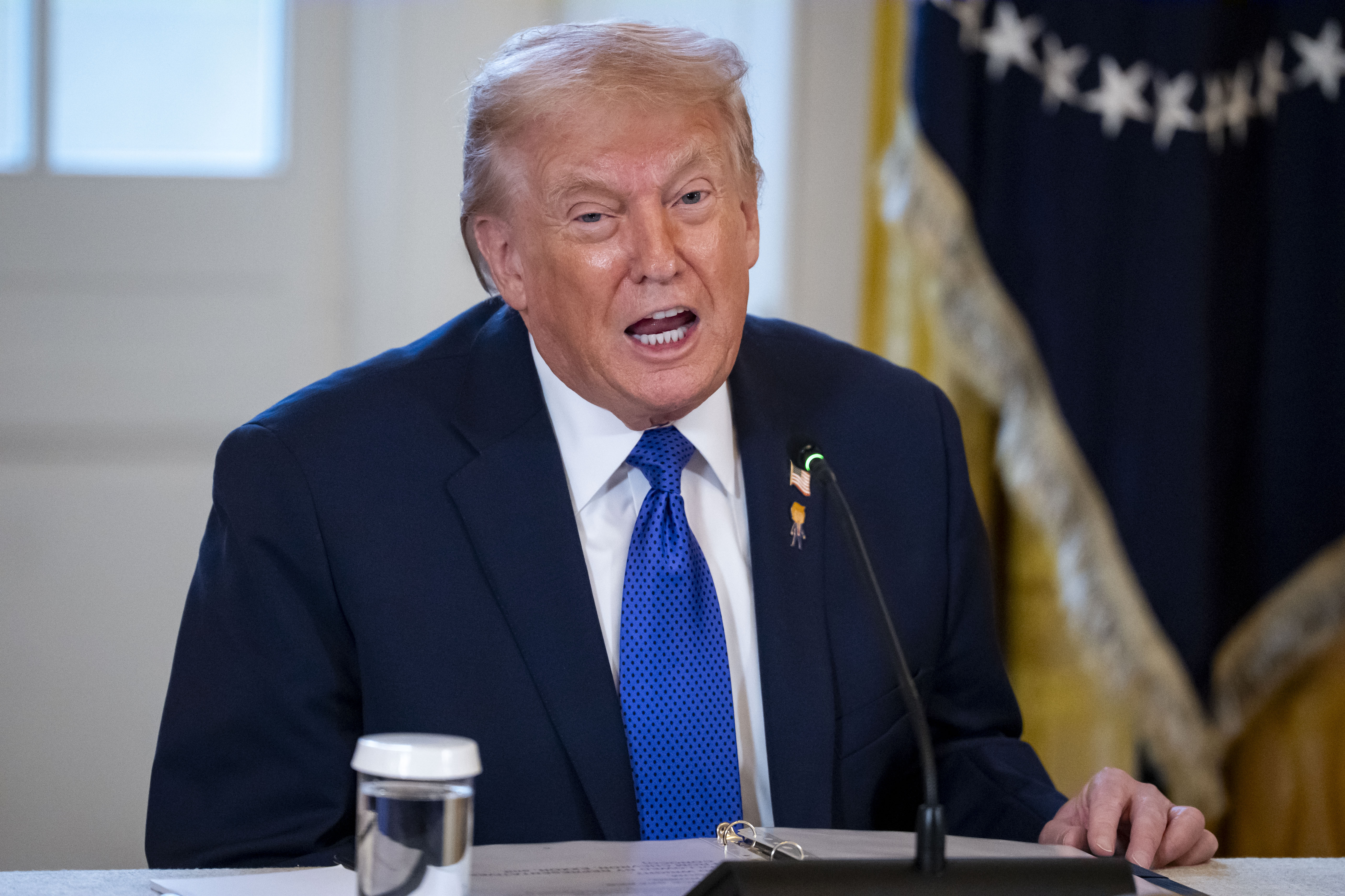Donald J. Trump speaking at a table with a microphone and water glass in front of him.