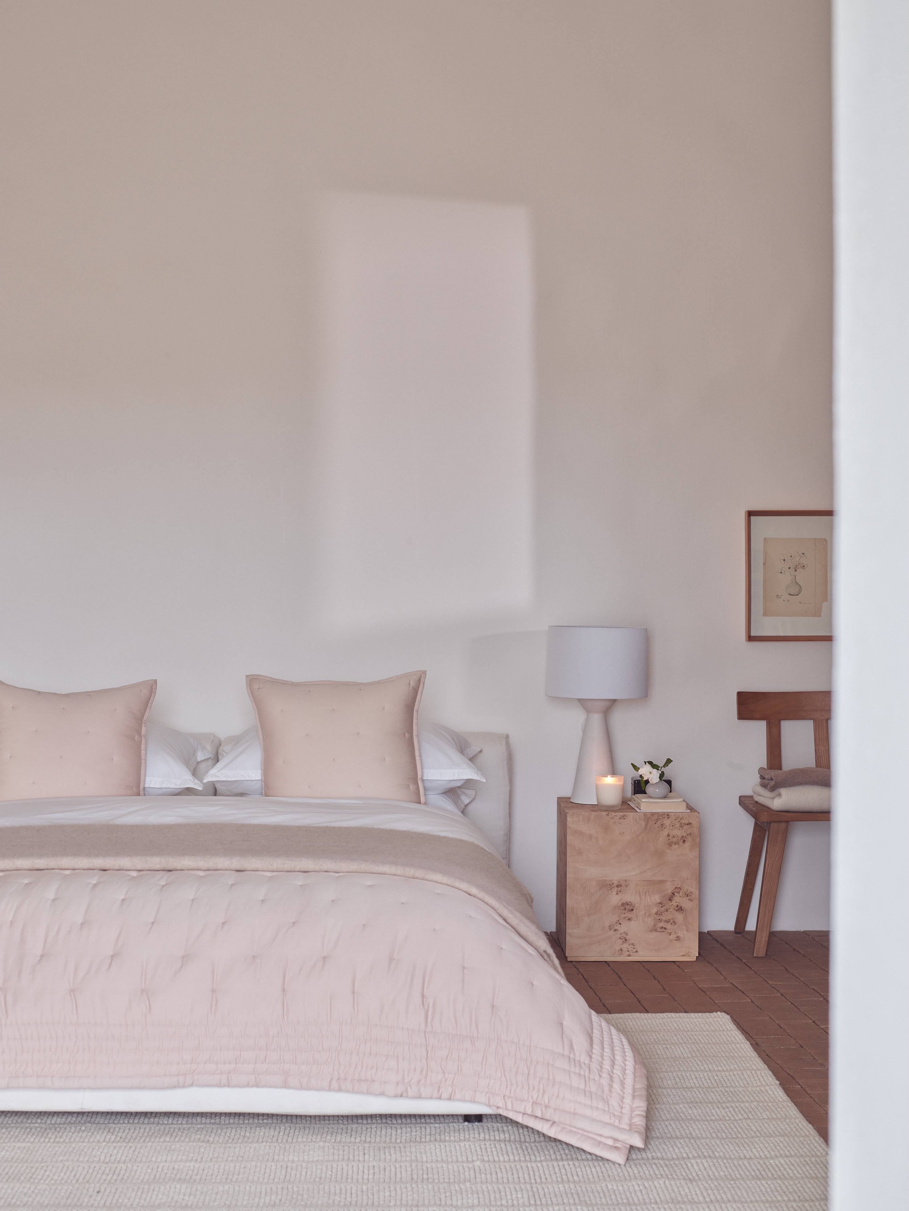 A bedroom with a pink quilted bedspread, a light wood nightstand with a lamp and candle, and a wooden chair with folded blankets.