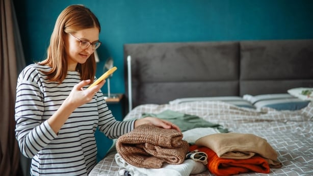 middle-aged woman in glasses photographs clothing laid out on a bed in a cozy bedroom using her smartphone, selling second-hand items and choosing clothes with the help of an online stylist at home.