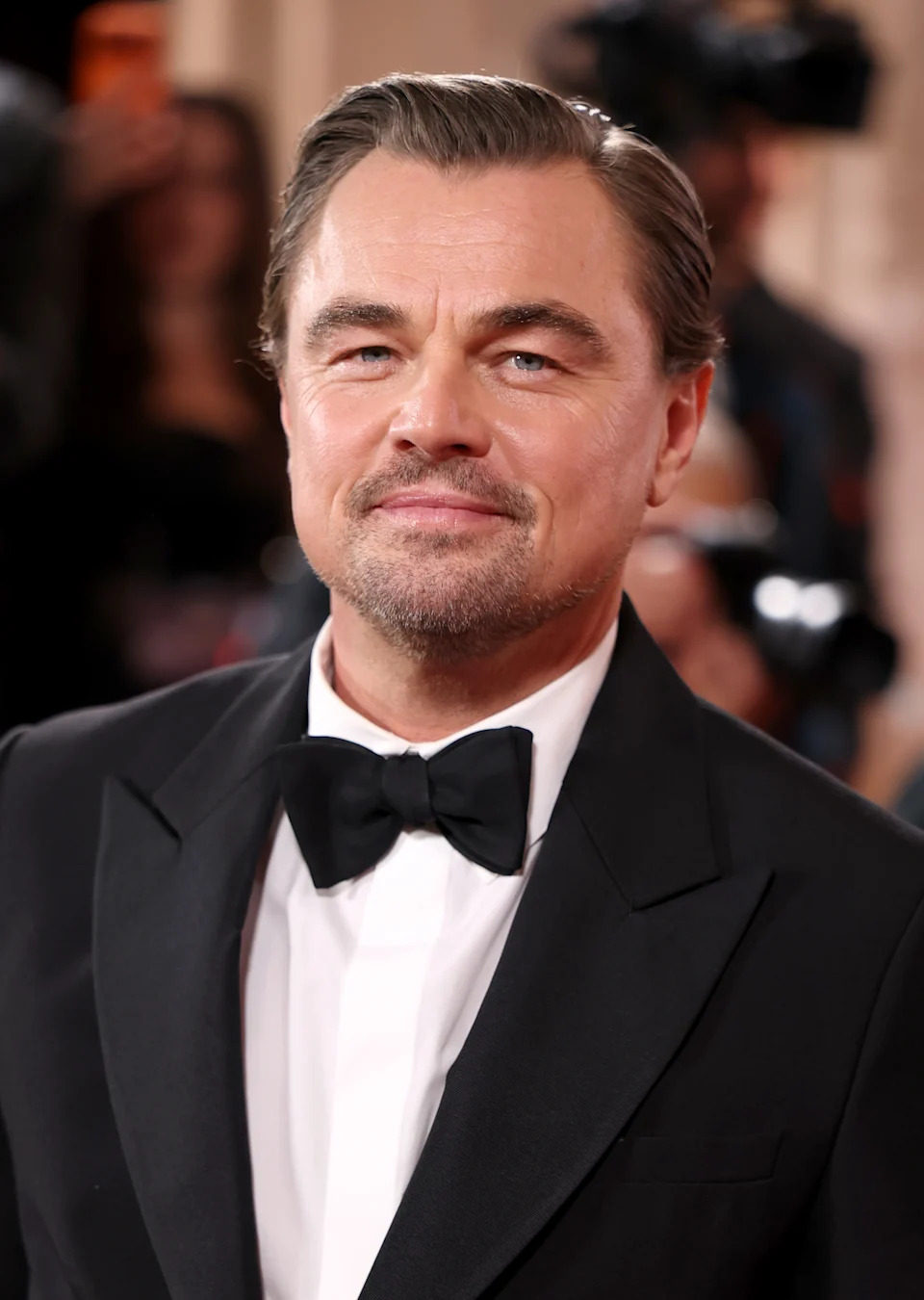 A person in a tuxedo poses on a red carpet, with people and cameras in the background