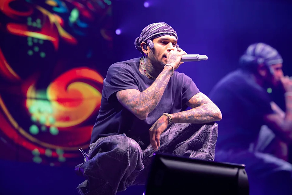 Chris Brown performing on stage, wearing a bandana and casual clothing, holding a microphone amidst vibrant, abstract background visuals