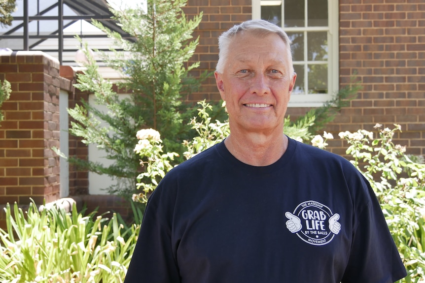A man in a blue shirt with a logo that says Grab Life by the Balls stands smiling in front of a garden 