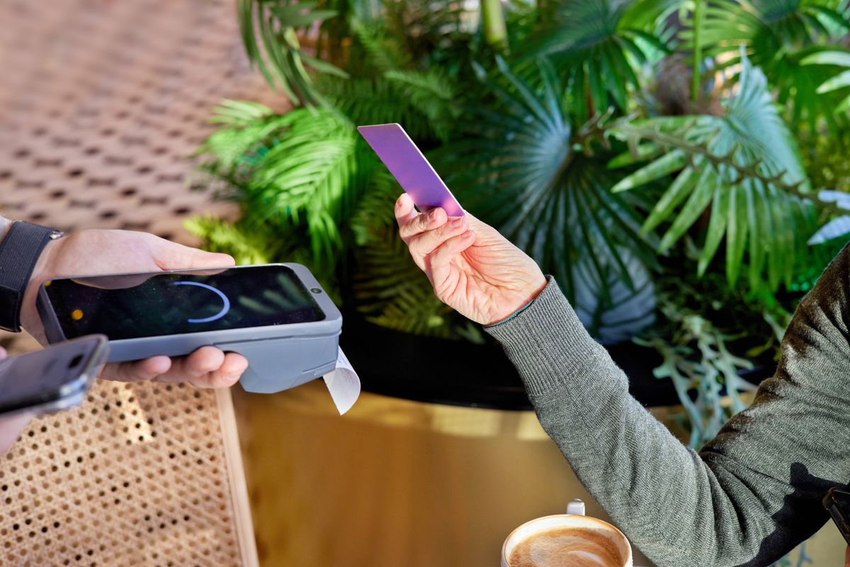 One person is making a contactless payment with a credit card at a point-of-sale terminal, completing a quick and secure transaction at a cafe with a warm beverage nearby