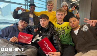 Four young men sit on a sofa with three young men standing behind them. Two of the four men who are seated are holding red cards saying 'North West Tonight'.