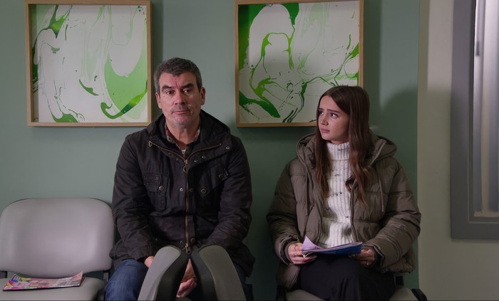 Sarah looks sideways towards Cain in a hospital in a scene from Emmerdale