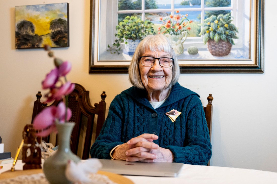 Ruth Gove smiles at her dining room table. She has a purple orchid on the table and some paintings of plants and nature on the wall behind her.