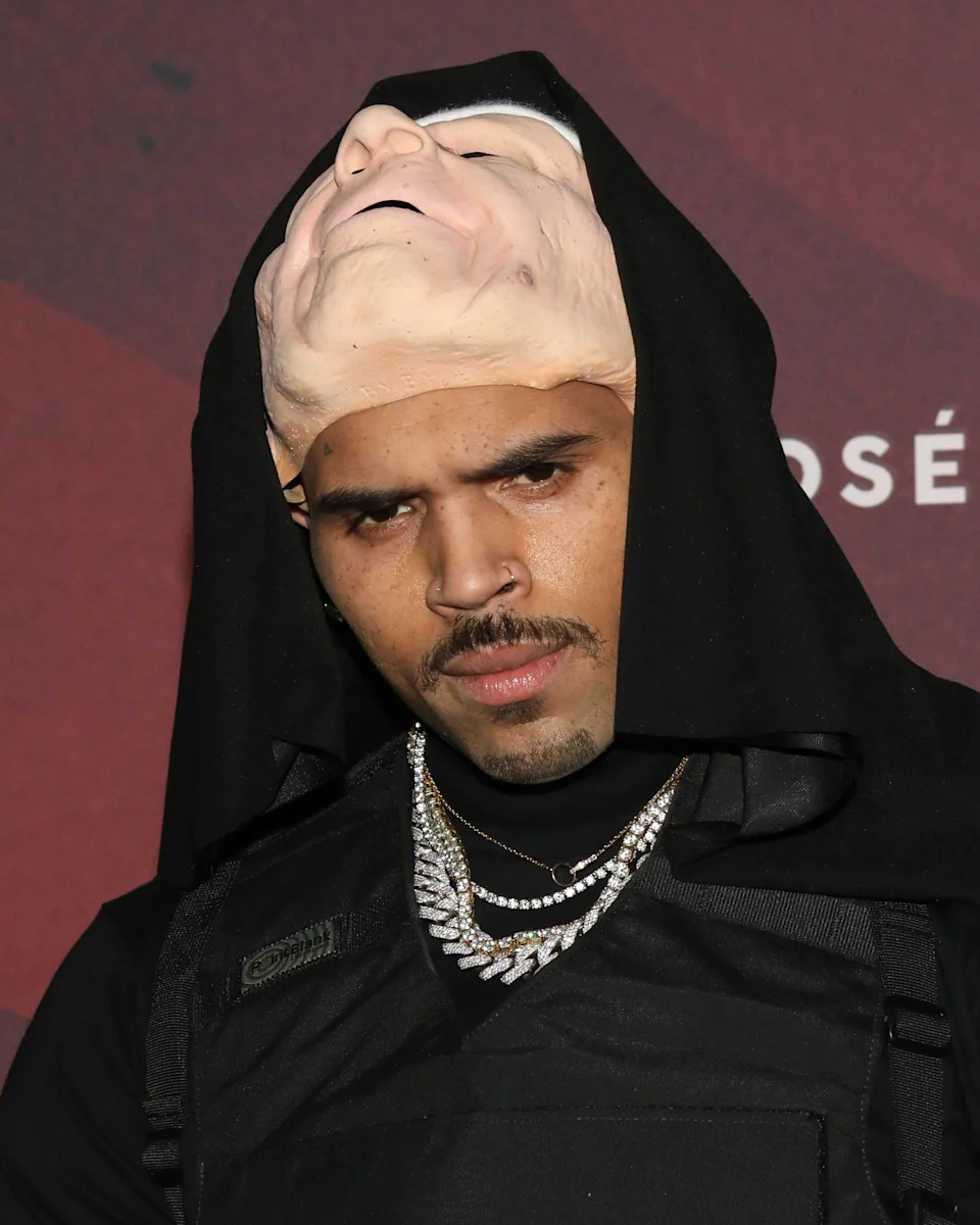 Chris Brown at a celebrity event wearing a mask over his head with layered necklaces and a vest