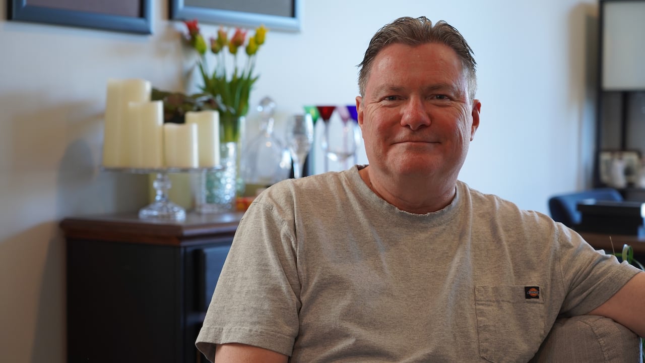 A man in a grey T-shirt sits in a living room. Behind him are tulips and wine glasses sitting on a counter.