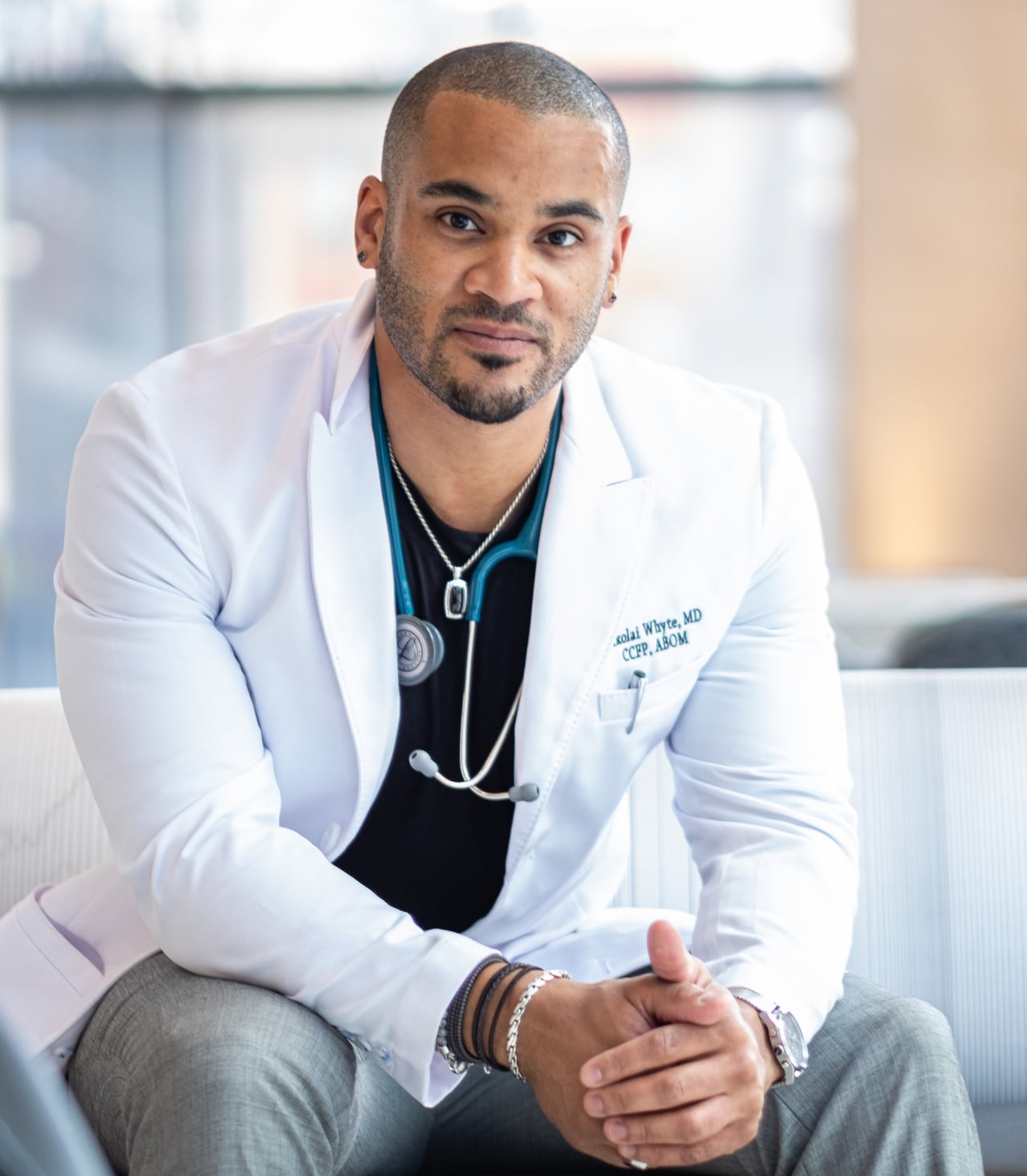 a Black man in a white physician coat and stethoscope sits and looks forward with hands clasped 