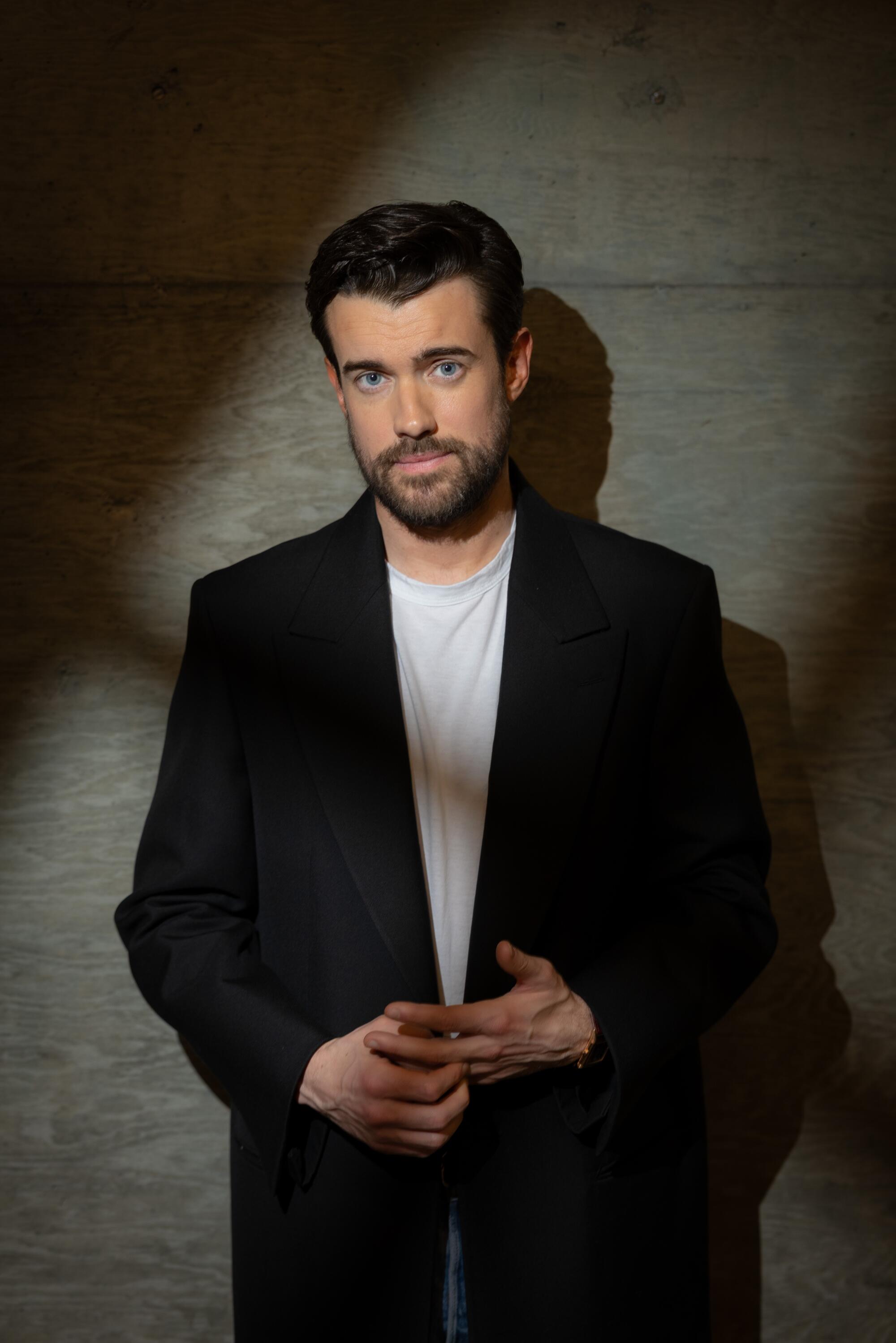 A man in a black blazer and white T-shirt leaning against a brick wall.