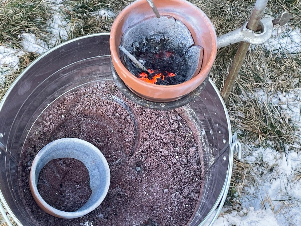 A close up of a metal bin with sand at bottom and a broken clay pot with something glowing inside,