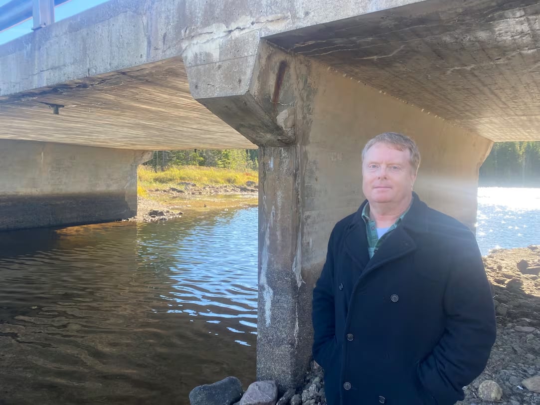 Man, standing near a low bridge with a stream nearby, looking at camera.