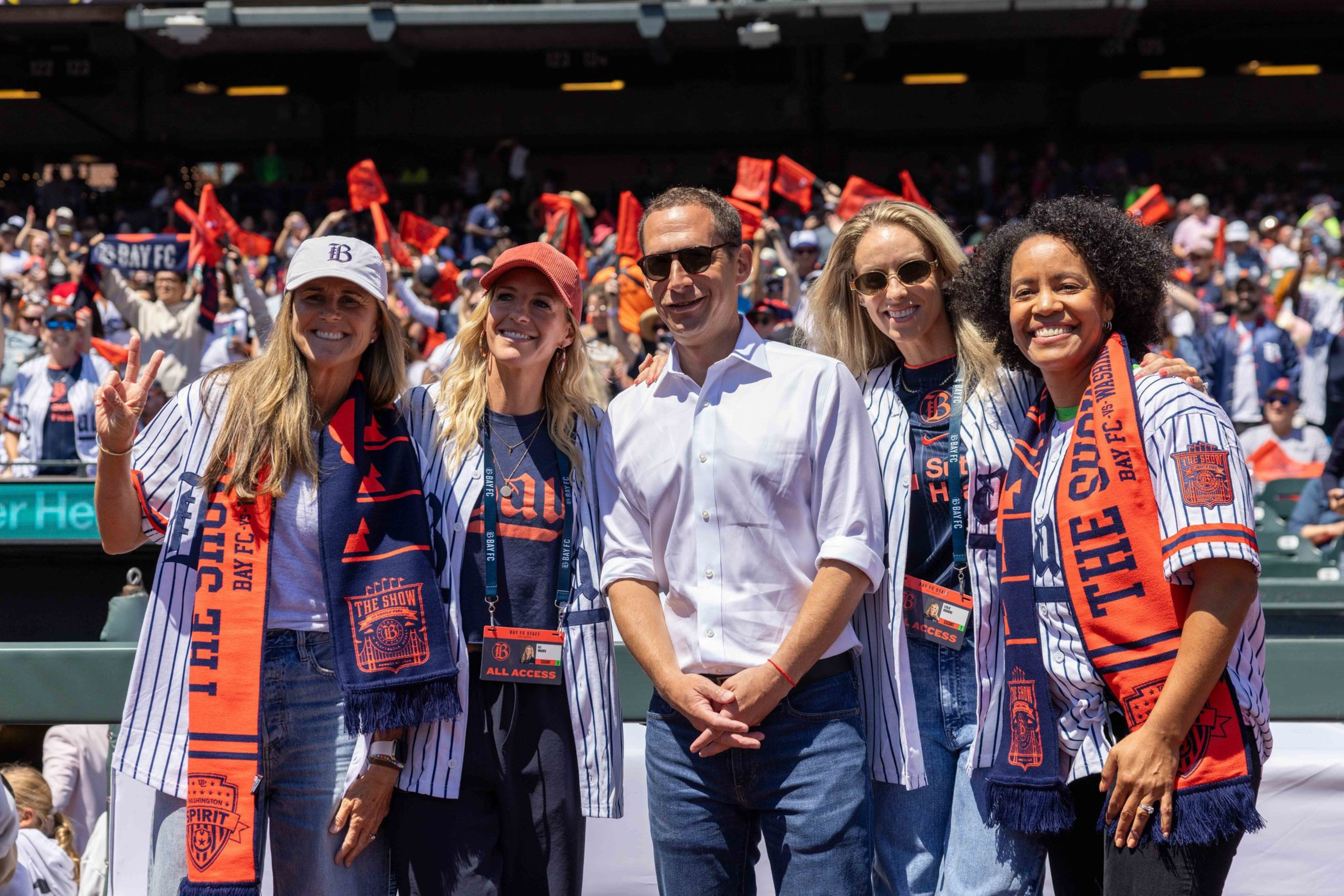 Five people, four women and one man, smile at the camera wearing baseball jerseys and scarves at a crowded stadium.