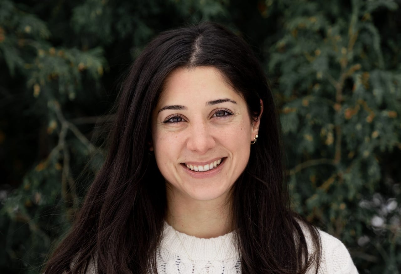 A woman with long, dark hair wearing a white, cable-knit sweater smiles while standing in front of a tree.