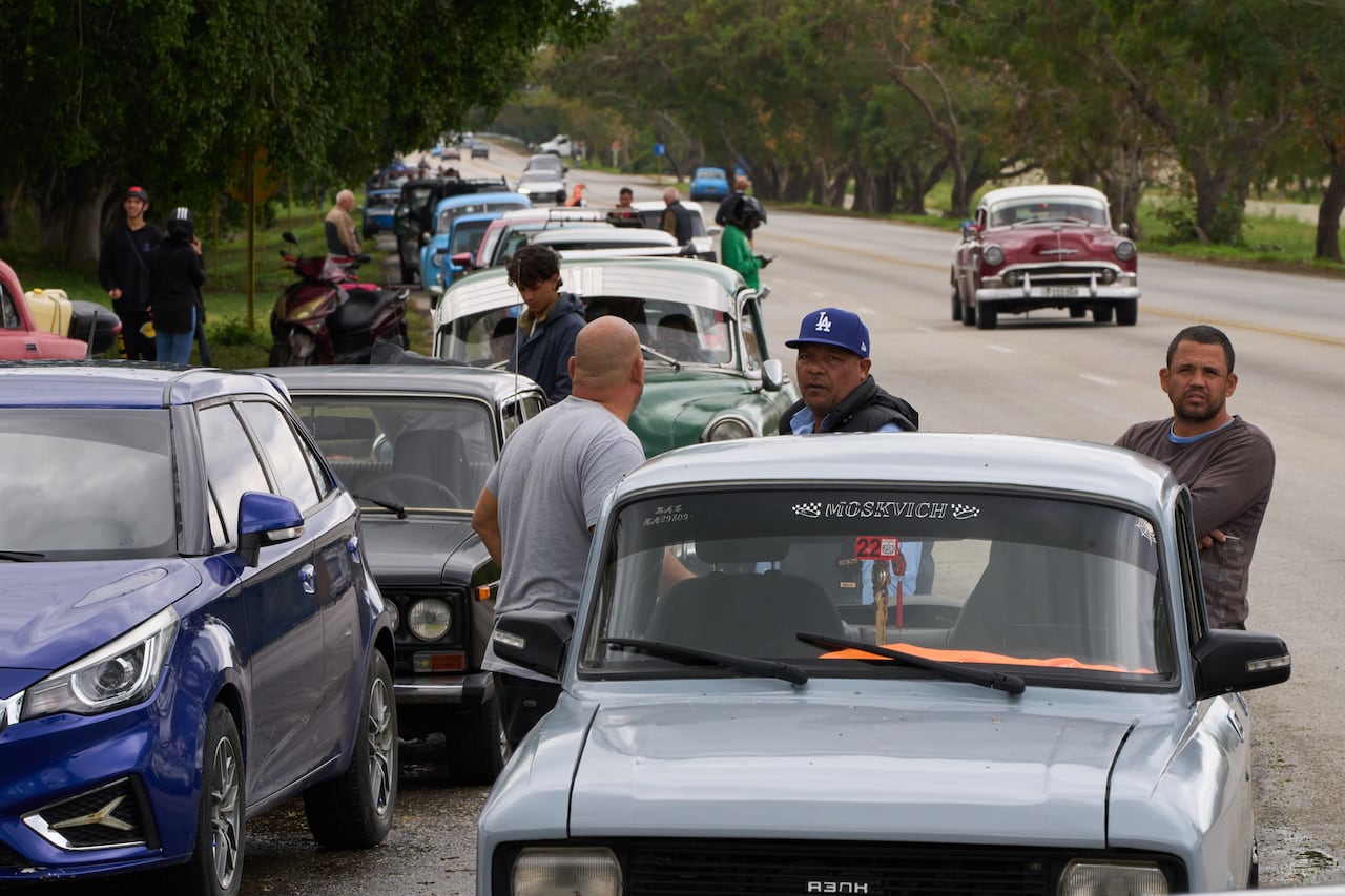 Several men stand outside an old car that's one of dozens waiting in a long lineup.