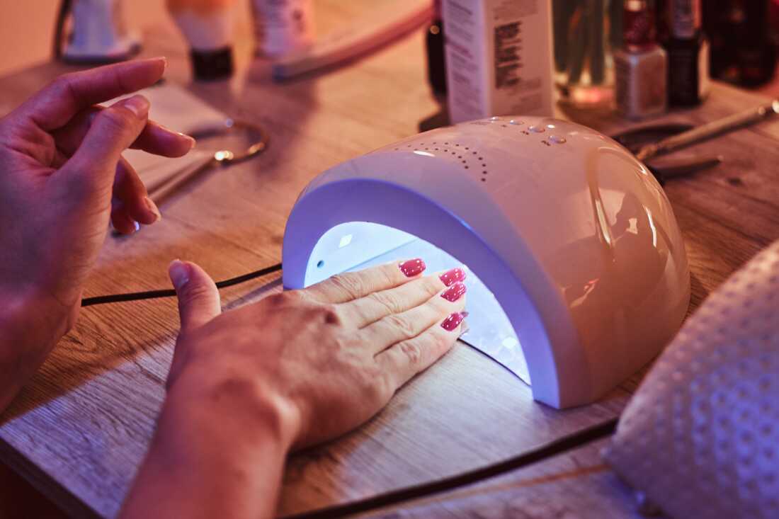 A client with red nail polish cures her gel manicure under a UV/LED nail lamp. 