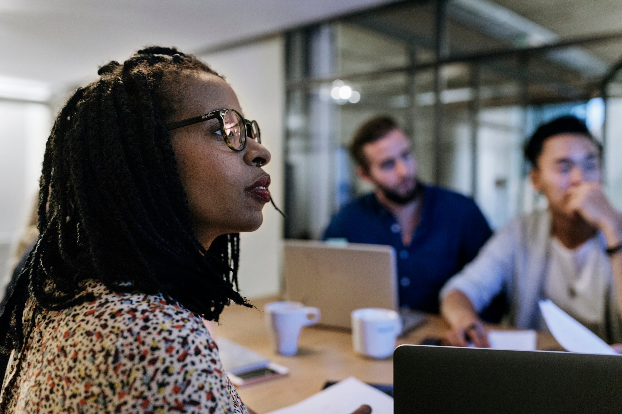 Three investors sit around a conference table with laptops and coffee cups while watching another investor present some data.