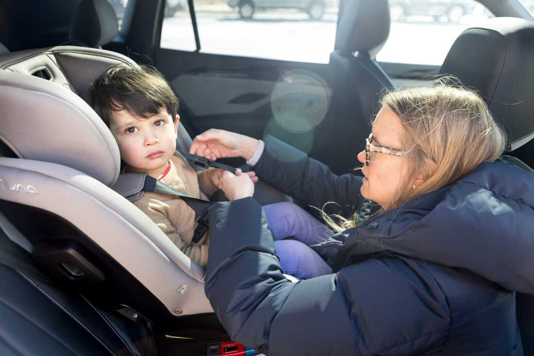A young boy sits in a car set in a car while Elizabeth Kanagawa demonstrates how to adjust the harness of the car seat.