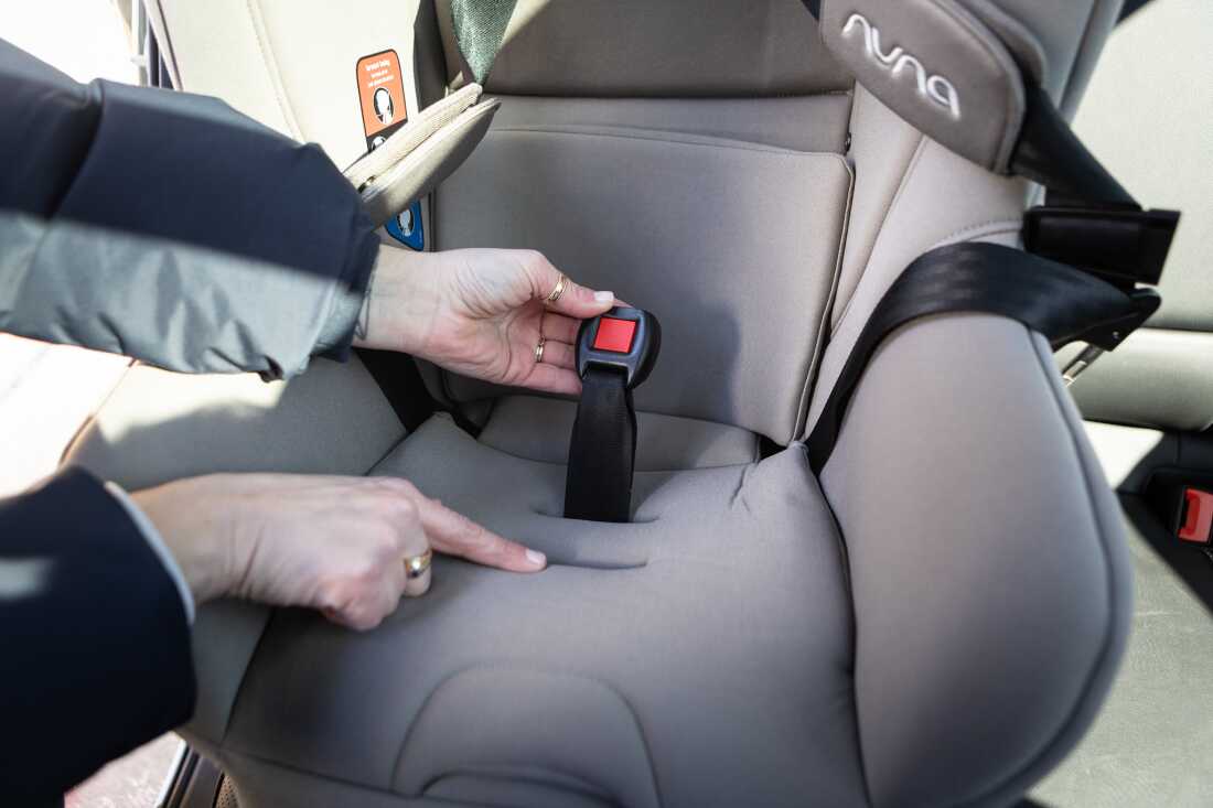 A person points out the crotch-buckle slots on a car seat, while holding the crotch buckle with one hand.