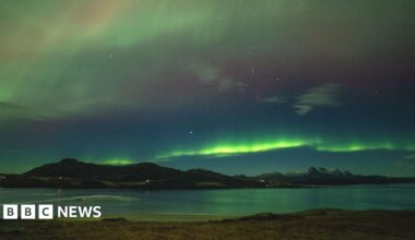 The Northern Lights in the north coast of Scotland. There are green and red stripes across the sky with snow-capped mountains below.