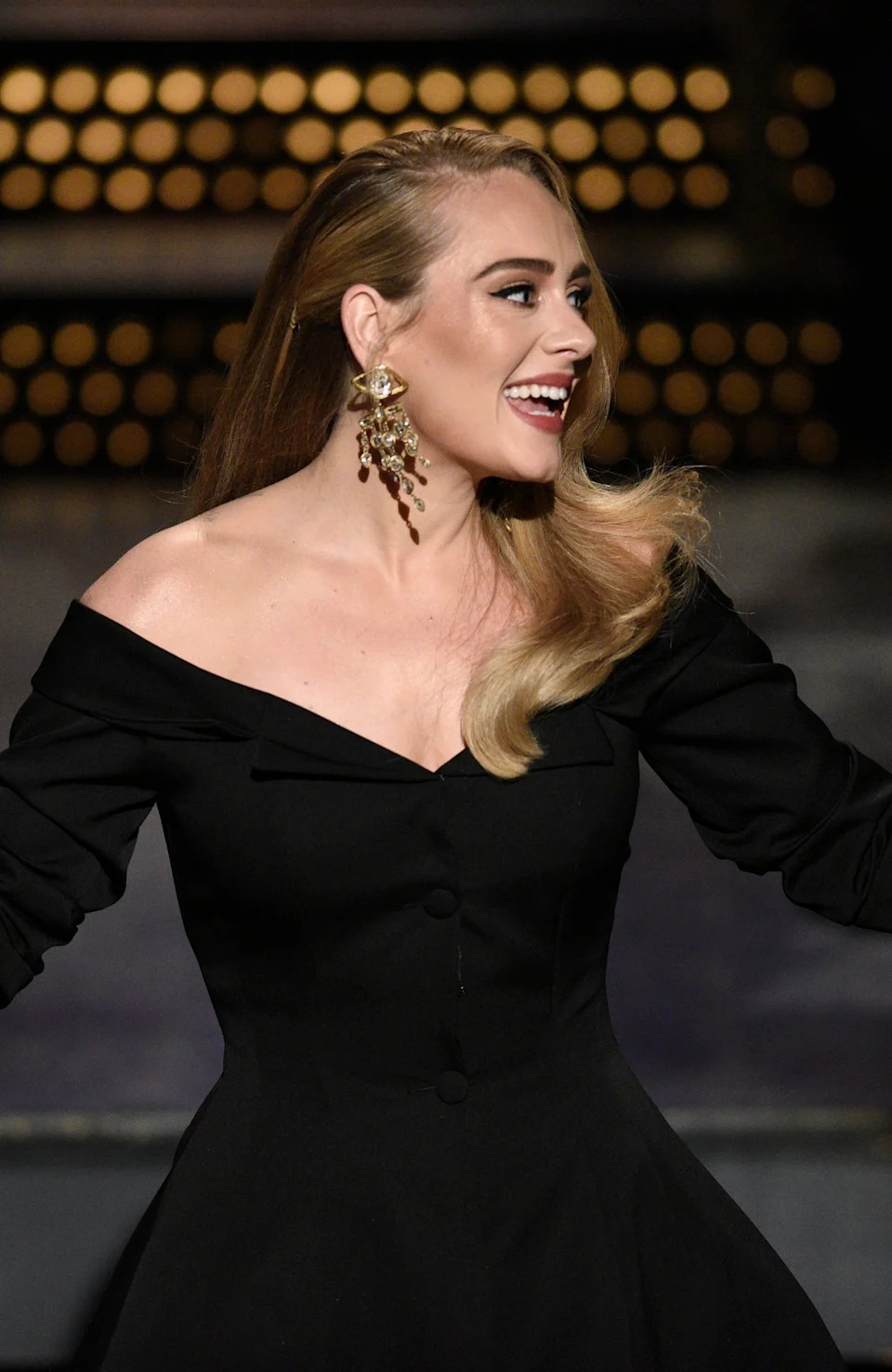 Person in elegant off-the-shoulder black dress and statement earrings, gesturing joyfully on stage