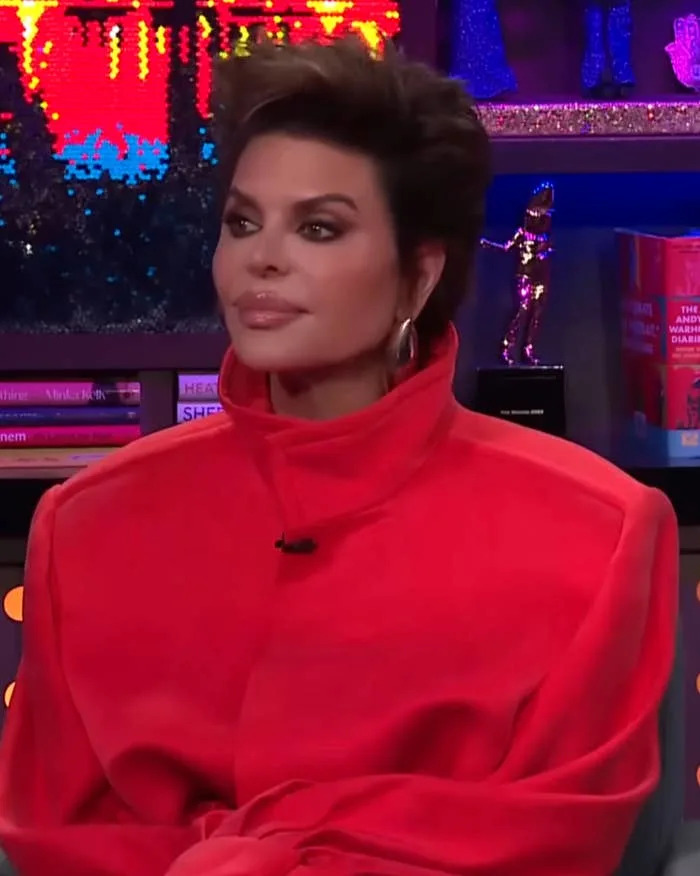 Person seated on talk show set, wearing a bold, structured outfit. Shelved books and decor are visible in the background