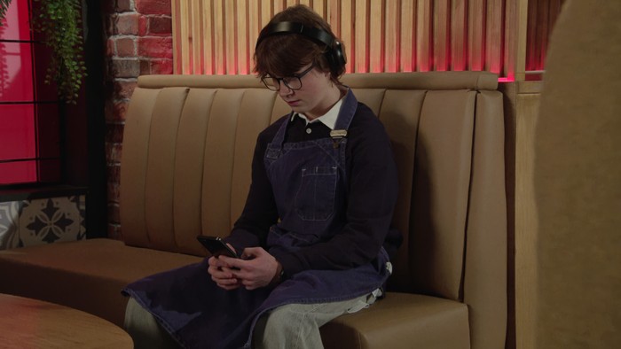 Sam sits on his phone in the bistro in a scene from Coronation Street