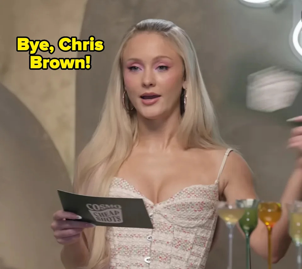 Zara Larsson with long blonde hair holding a card, standing near cocktail glasses. Wearing a white, textured top with thin straps