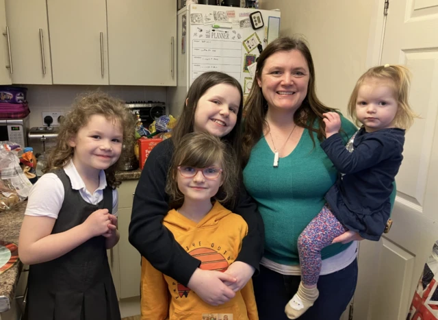 Patricia with her four daughters in the kitchen at home. Three of the girls stand next to her while she holds the youngest in her arm. They are looking at the camera and smiling