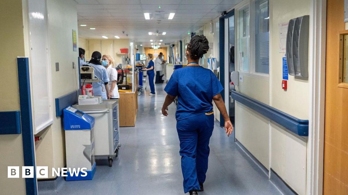 A nurse wears blue scrubs as she walks down a hospital corridor, there are other nurses in the background doing different tasks.