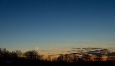 A orange and blue night sky with a glowing crescent moon and a bright dot for the planet Mercury with a series of silhouettes of trees in the foreground