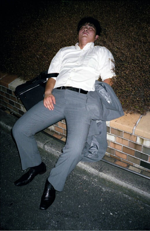 A man in a white shirt and gray suit pants is slumped asleep on a low brick wall at night, with his jacket and briefcase beside him and his mouth slightly open.