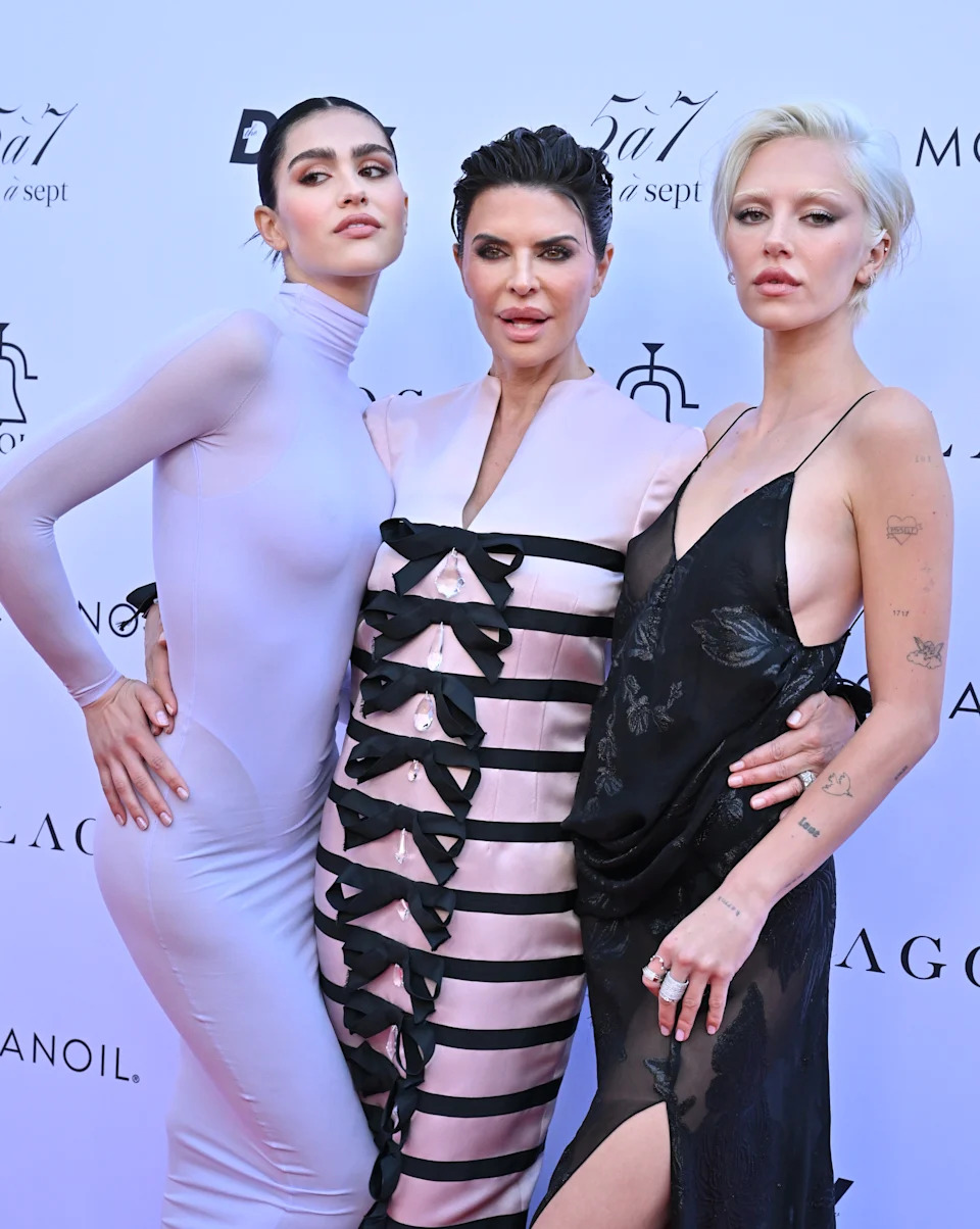 Three individuals pose on the red carpet. The center person wears a dress with bow details, flanked by others in form-fitting and slip dresses