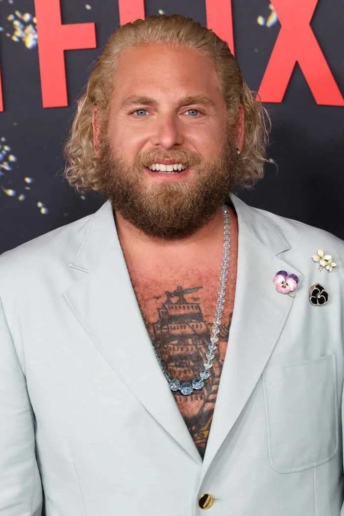Person with curly hair and beard on the red carpet, wearing a light blazer adorned with flower pins, revealing a tattooed chest