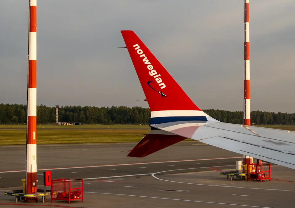Airplane wing with "Norwegian" text on the tarmac, surrounded by landing lights at an airport, under a partly cloudy sky