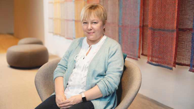 A woman with short blonde hair sits with her hands folded in a chair and a half-smile, with reddish fabrics hanging behind her.