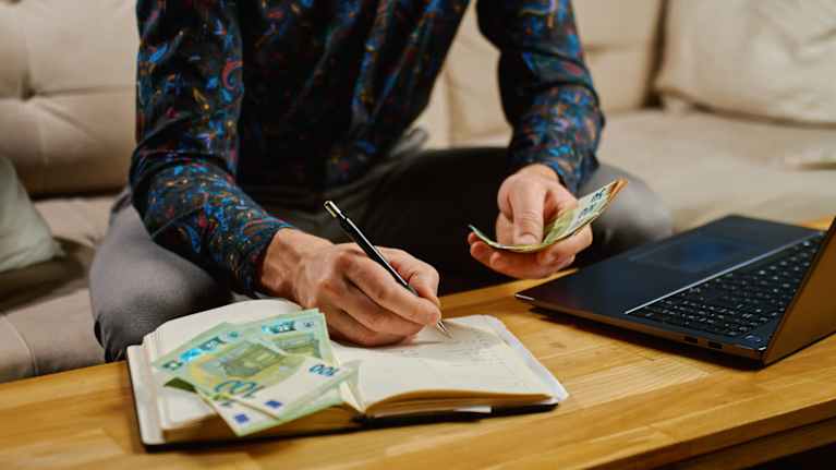 A man sits by a laptop and notebook counting money.