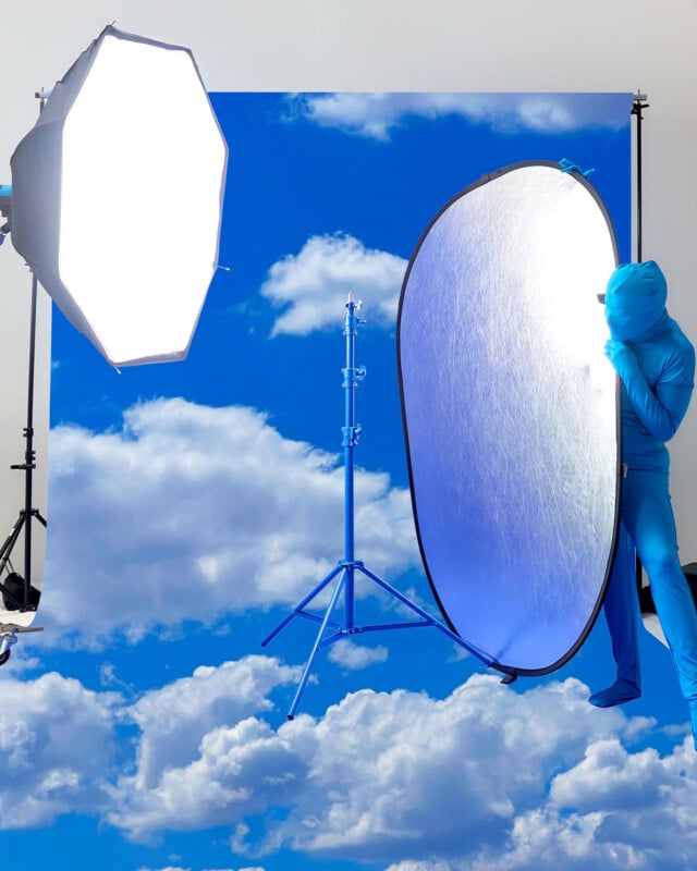 A person in a blue bodysuit holds a large reflector in front of a cloud backdrop, with bright studio lights and photography equipment visible in the scene.