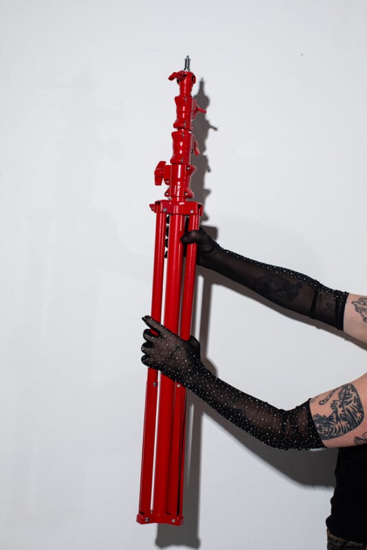 A person wearing black, sheer, rhinestone-studded gloves holds a folded, bright red tripod against a white wall. Their tattooed arms are partially visible.