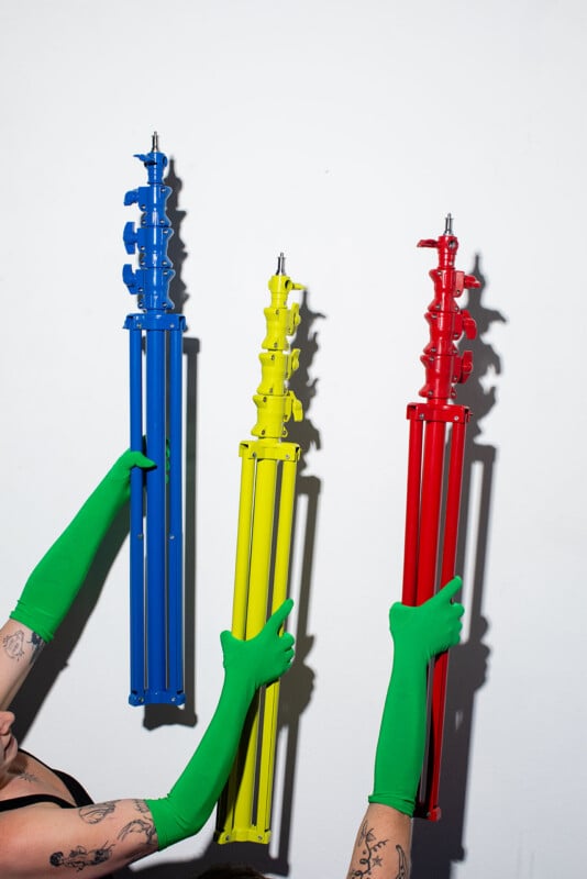 Three hands wearing bright green gloves hold up three tripod stands, colored blue, yellow, and red, against a plain white background. Two arms have visible tattoos.