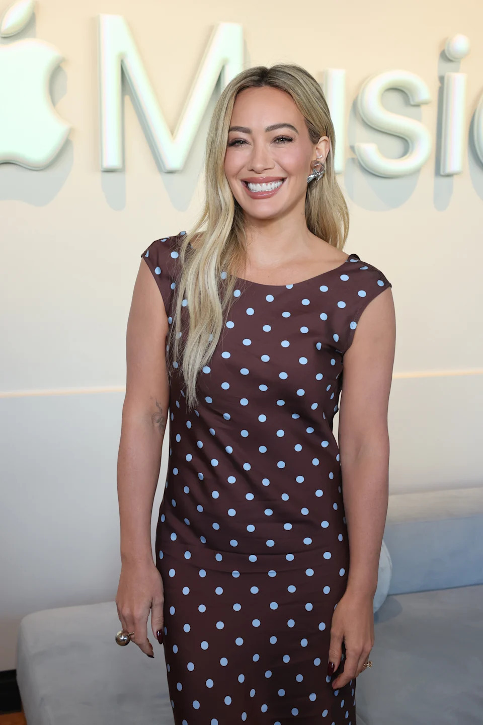 A person smiles on a red carpet, wearing a fitted brown dress with white polka dots, standing in front of an Apple Music backdrop