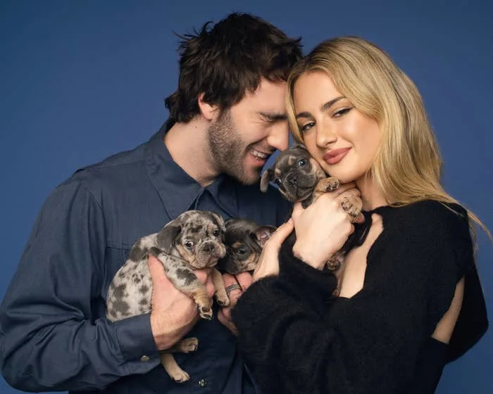 A couple joyfully holds three puppies, each with a unique coat. The man wears a button-up shirt; the woman is in a cozy sweater