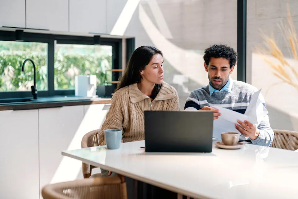 Two people discussing documents at a kitchen table with a laptop and coffee cups, conveying a work-related scenario
