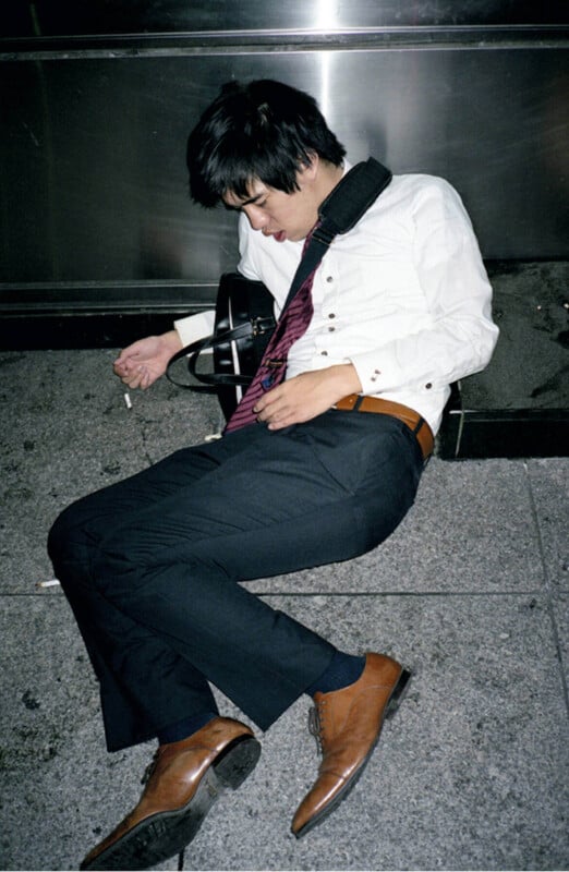 A man in dress clothes and brown shoes sleeps on a city sidewalk, leaning against a metal wall with a shoulder bag and a cigarette in his hand.