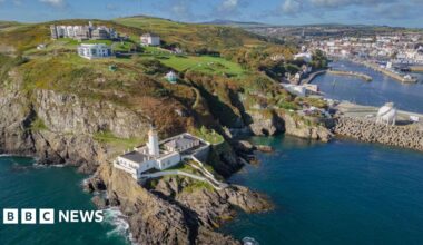 An arial view of Douglas Head, you can see the lighthouse on a rocky terrain, there are buildings scattered on the green terrain.